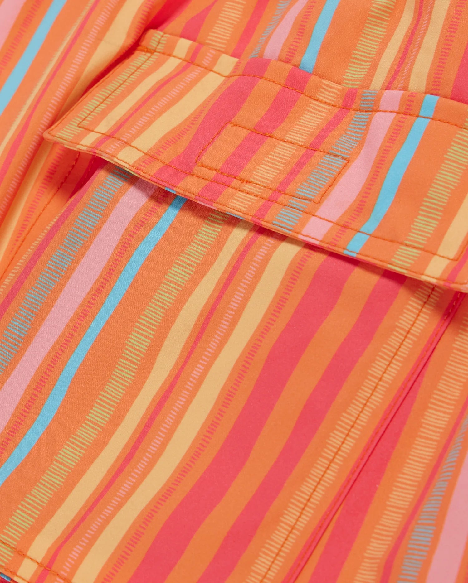 Close-up of a brightly colored fabric with a pattern of vertical stripes in various shades of orange, pink, and blue. The fabric appears to be part of MENS ANTON ALL OVER PRINT SWIM TRUNK - B6W542C200 by Psycho Bunny, with a visible pocket and stitching details.
