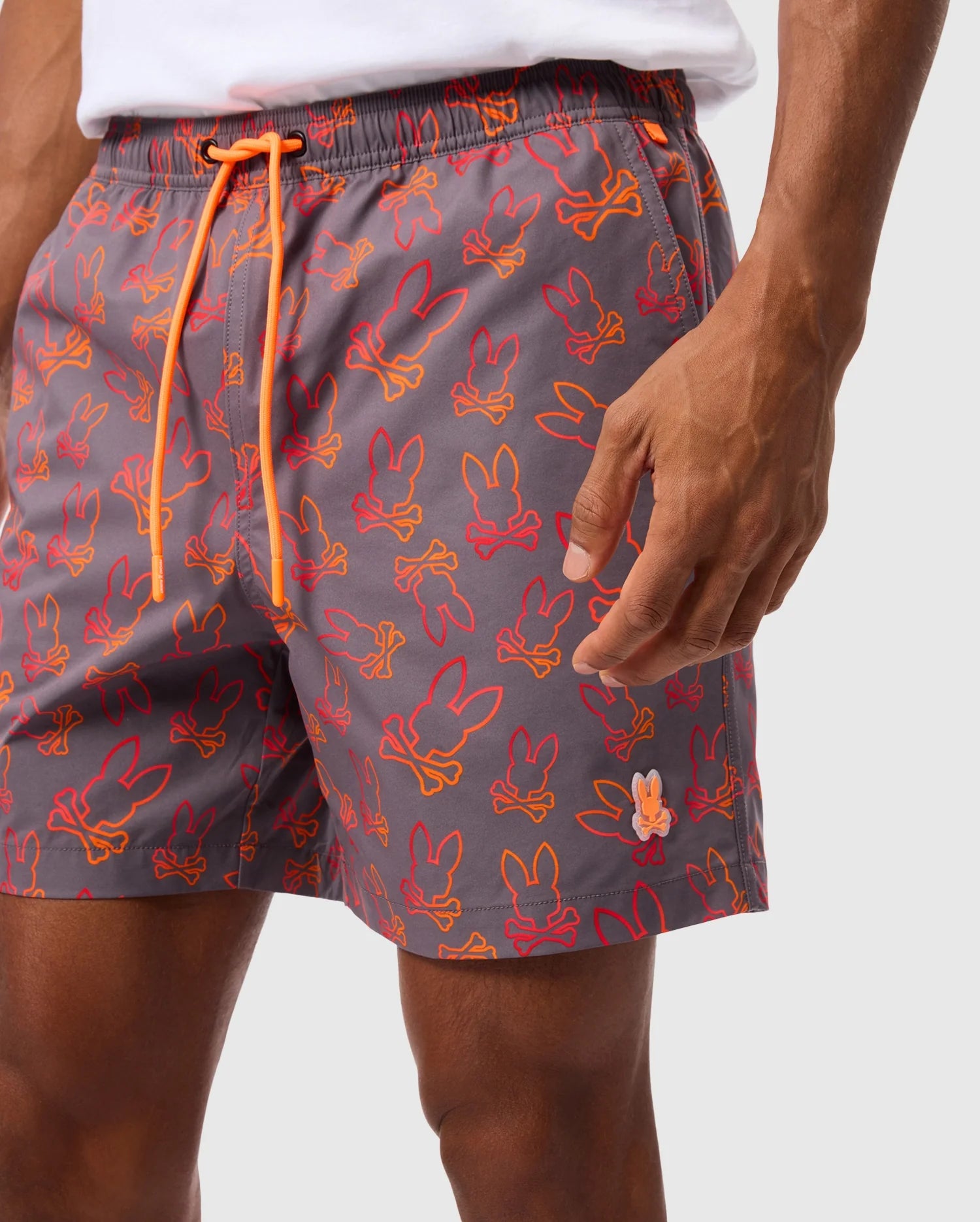 A person is shown from mid-torso to mid-thigh, wearing Psycho Bunny's MENS SHELDON ALL OVER PRINT SWIM TRUNK (Product Code: B6W588C200), featuring a gray base with orange and red bunny silhouettes. The swim trunks include an orange drawstring and are crafted from quick-dry fabric. The person is also wearing a white shirt against a plain white background.