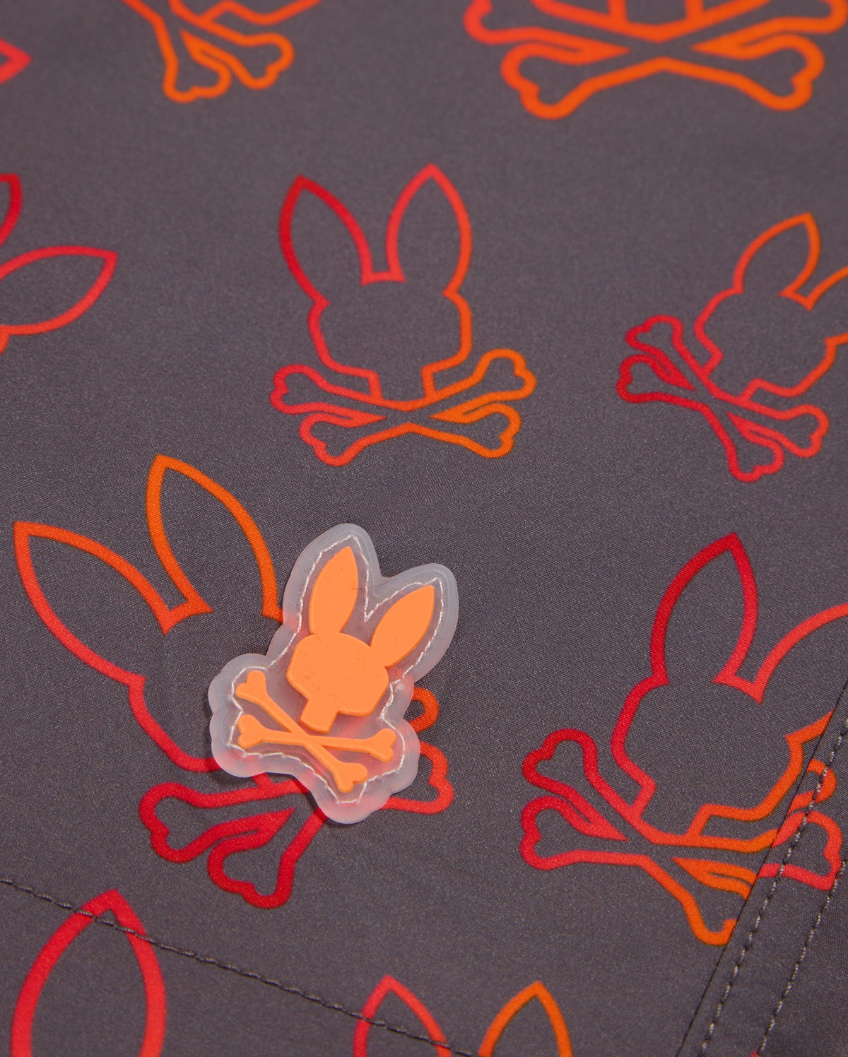 A close-up of the MENS SHELDON ALL OVER PRINT SWIM TRUNK - B6W588C200 by Psycho Bunny showcases a repeating bunny pattern with silhouettes and crossed bones, printed in vibrant red and orange on a dark grey quick-dry fabric. A small rubber patch with the same design is sewn onto the material.