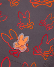 A close-up of the MENS SHELDON ALL OVER PRINT SWIM TRUNK - B6W588C200 by Psycho Bunny showcases a repeating bunny pattern with silhouettes and crossed bones, printed in vibrant red and orange on a dark grey quick-dry fabric. A small rubber patch with the same design is sewn onto the material.