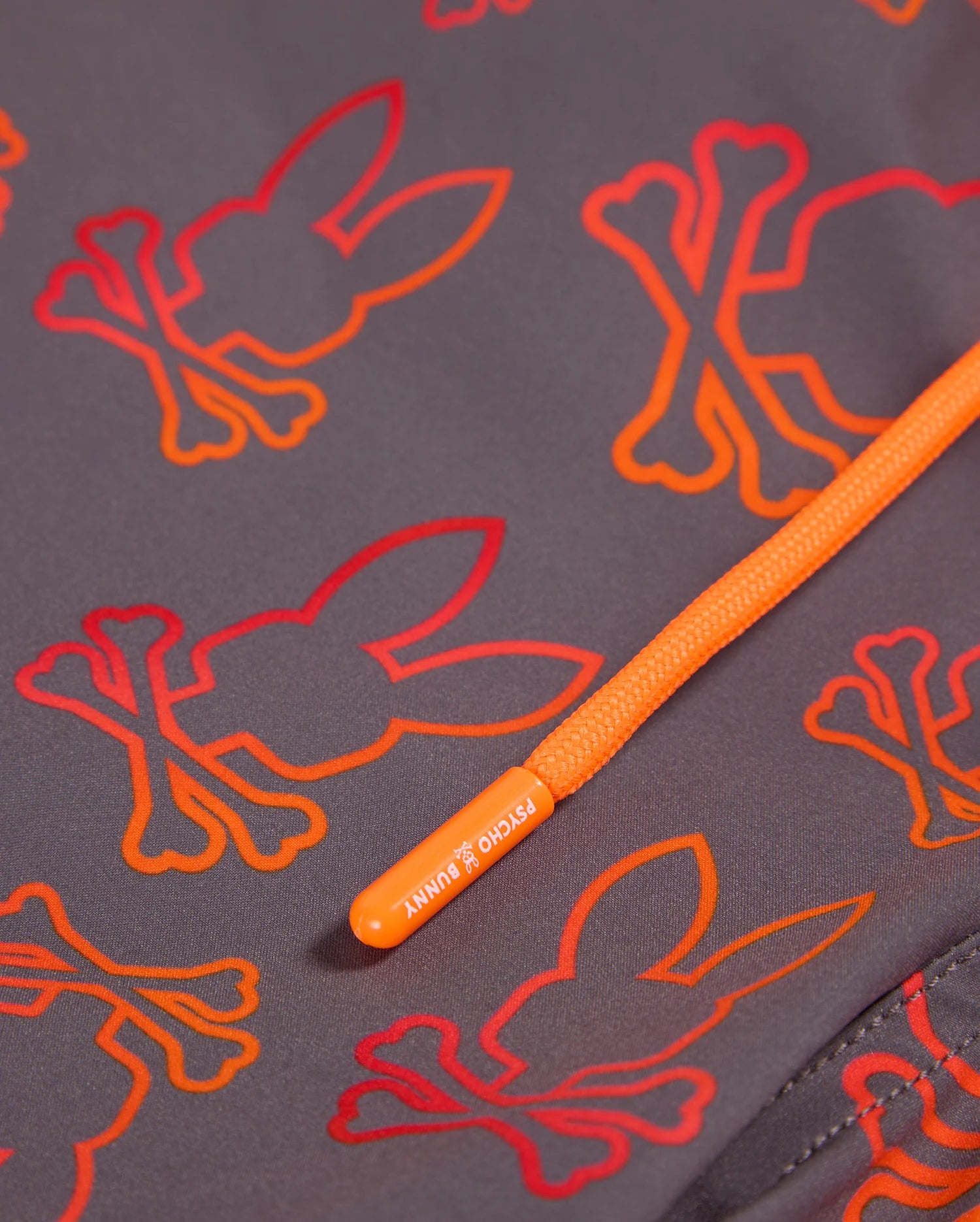 Close-up of the Psycho Bunny Men's Sheldon All Over Print Swim Trunk - B6W588C200, showcasing an intricate bunny pattern with orange and red bunny skull-and-crossbones designs. An orange drawstring with a branded tip labeled "PSYCHO BUNNY" crosses diagonally over the quick-dry grey fabric.
