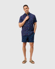 A person stands against a plain background, wearing navy blue short-sleeve button-up shirt and **MENS SHELDON ALL OVER PRINT SWIM TRUNK - B6W588C200** adorned with a quirky bunny pattern by **Psycho Bunny**. Made of quick-dry fabric, the outfit is complemented by tan sandals. One hand adjusts the shirt collar while the other rests in a pocket, looking confidently at the camera.