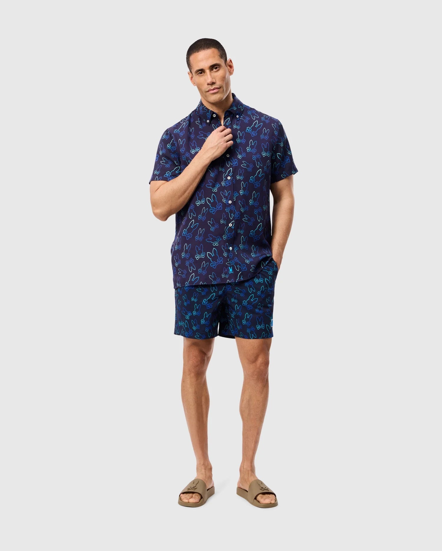 A person stands against a plain background, wearing navy blue short-sleeve button-up shirt and **MENS SHELDON ALL OVER PRINT SWIM TRUNK - B6W588C200** adorned with a quirky bunny pattern by **Psycho Bunny**. Made of quick-dry fabric, the outfit is complemented by tan sandals. One hand adjusts the shirt collar while the other rests in a pocket, looking confidently at the camera.
