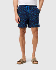 A man is standing against a plain background, wearing Psycho Bunny MENS SHELDON ALL OVER PRINT SWIM TRUNK - B6W588C200. He is also wearing a white T-shirt and beige slides. His hands are in the pockets of his swim trunks. Only the lower half of his body is visible.
