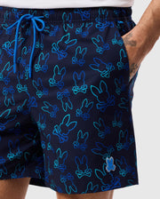 A person wearing Psycho Bunny MENS SHELDON ALL OVER PRINT SWIM TRUNK - B6W588C200 featuring a repeating bunny pattern in blue and green outlines. The quick-dry fabric shorts have a blue drawstring, and the person's right hand is in their pocket. A small tattoo is visible on their left wrist.