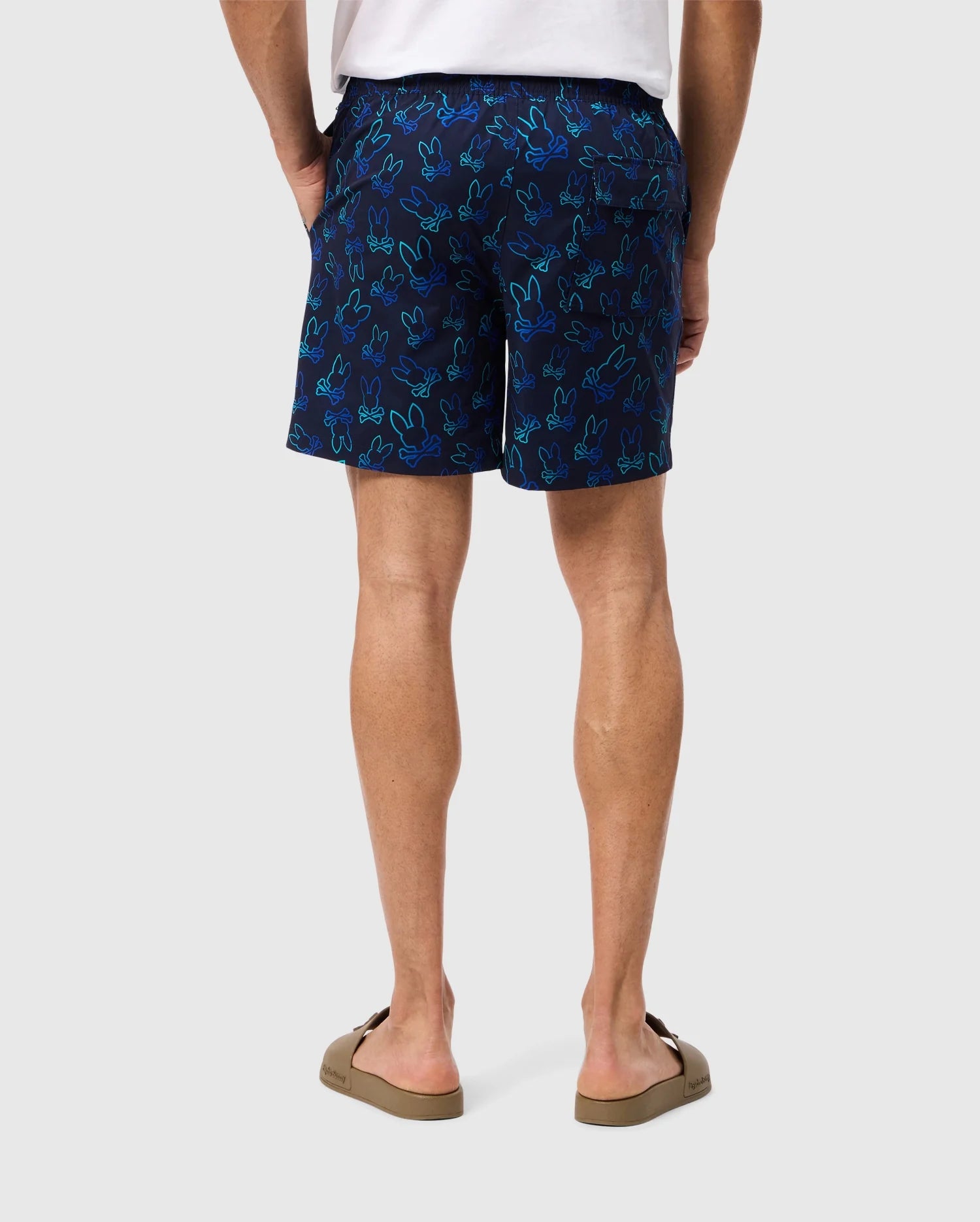 The image shows a person from the waist down, wearing dark blue Psycho Bunny MENS SHELDON ALL OVER PRINT SWIM TRUNK - B6W588C200 with a light blue bunny pattern. The person is also wearing brown flip-flops and a white shirt. The background is plain and neutral.