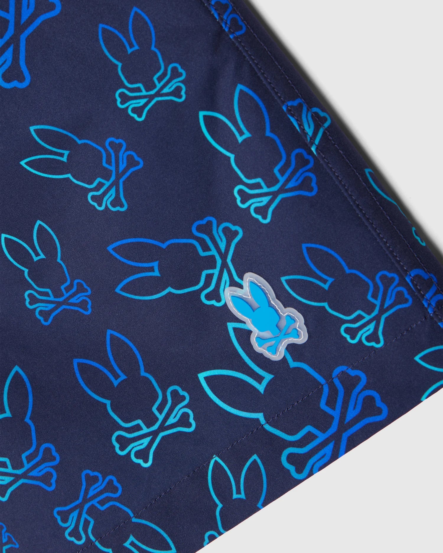 Close-up of MENS SHELDON ALL OVER PRINT SWIM TRUNK - B6W588C200 by Psycho Bunny, made from quick-dry fabric, featuring a repetitive bunny pattern: turquoise outline bunny heads with crossbones underneath. A smaller, solid blue bunny logo is also visible on the navy blue fabric.