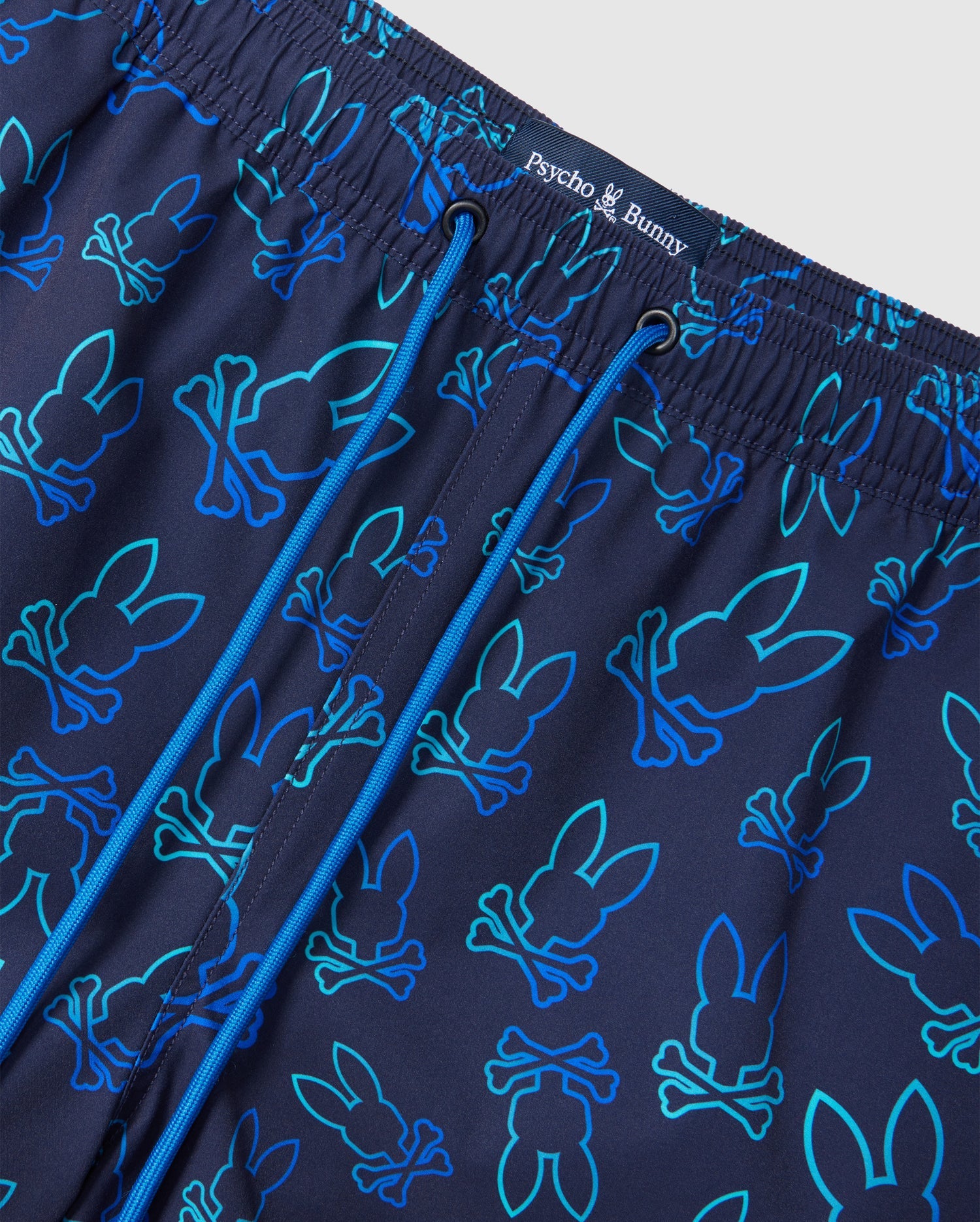 Close-up of the MENS SHELDON ALL OVER PRINT SWIM TRUNK - B6W588C200 featuring an all-over Bunny pattern in various shades of blue and teal. The waistband has an elastic band with blue drawstrings and the brand name "Psycho Bunny" printed at the center.