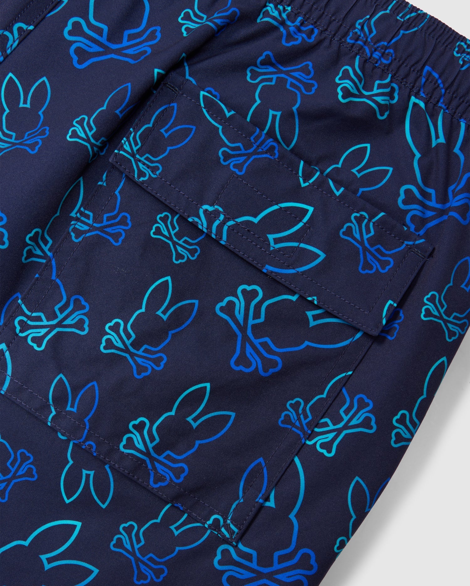 Close-up of a pair of dark blue MENS SHELDON ALL OVER PRINT SWIM TRUNK - B6W588C200 by Psycho Bunny with an elastic waistband. The shorts feature an all-over bunny pattern with crossbones in shades of teal and blue. Made from quick-dry fabric, they also have a patch pocket with a flap, showcasing the playful print pattern.