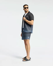 A man confidently models Psycho Bunny's MENS BENDIGO ALL OVER PRINTED SWIM TRUNK (B6W619F200) with a matching patterned shirt, black sandals, and sunglasses. Striking a cool pose against a white background, he offers a stylish swimwear upgrade.