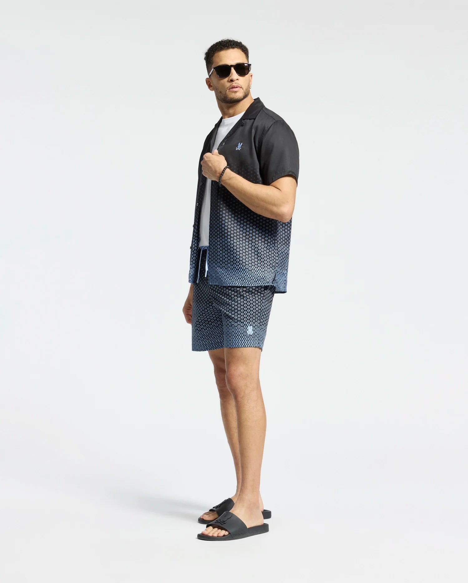 A man confidently models Psycho Bunny's MENS BENDIGO ALL OVER PRINTED SWIM TRUNK (B6W619F200) with a matching patterned shirt, black sandals, and sunglasses. Striking a cool pose against a white background, he offers a stylish swimwear upgrade.