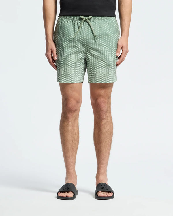 A person wears Psycho Bunny's MENS BENDIGO PRINTED SWIM TRUNK (B6W619F200) with an elasticized waist and drawstring, along with black slide sandals. Made of quick-dry fabric, these light green patterned shorts provide both style and function. Upper body is cropped against a white background.
