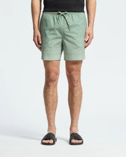 A person wears Psycho Bunny's MENS BENDIGO PRINTED SWIM TRUNK (B6W619F200) with an elasticized waist and drawstring, along with black slide sandals. Made of quick-dry fabric, these light green patterned shorts provide both style and function. Upper body is cropped against a white background.