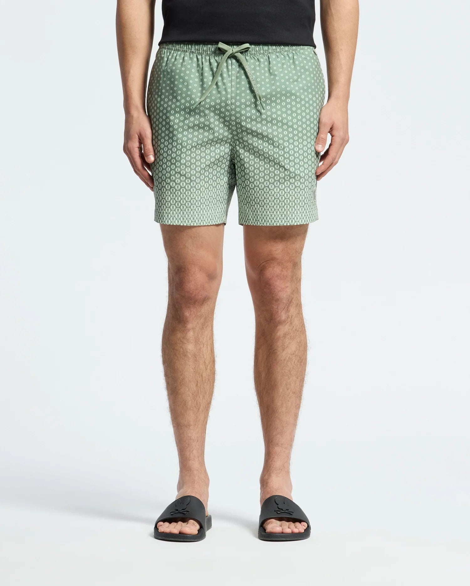 A person wears Psycho Bunny's MENS BENDIGO PRINTED SWIM TRUNK (B6W619F200) with an elasticized waist and drawstring, along with black slide sandals. Made of quick-dry fabric, these light green patterned shorts provide both style and function. Upper body is cropped against a white background.