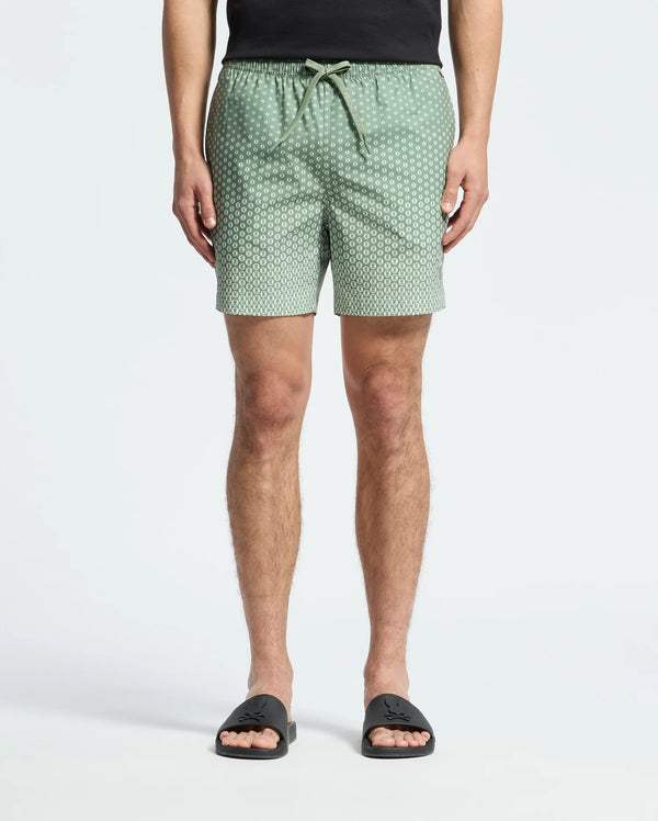 A person wears Psycho Bunny's MENS BENDIGO PRINTED SWIM TRUNK (B6W619F200) with an elasticized waist and drawstring, along with black slide sandals. Made of quick-dry fabric, these light green patterned shorts provide both style and function. Upper body is cropped against a white background.