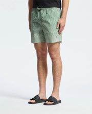 Someone is wearing Psycho Bunny's MENS BENDIGO PRINTED SWIM TRUNK - B6W619F200, featuring a light green pattern and elasticized waist. Paired with black sandals, the quick-dry fabric adds comfort. The upper body is partially visible against a simple light-colored background.