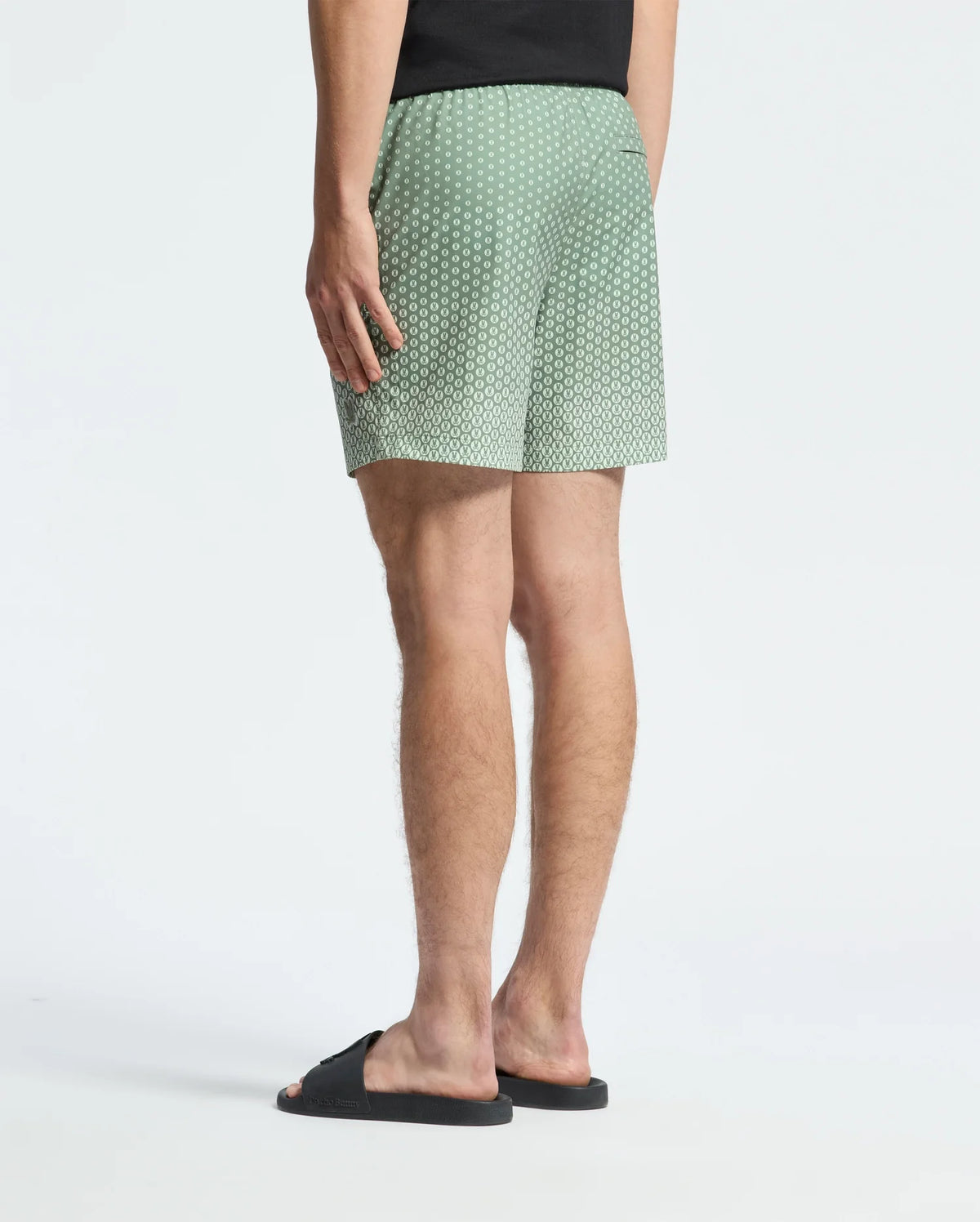 A person wearing Psycho Bunny's MENS BENDIGO PRINTED SWIM TRUNK in green with white dots, an elasticized waist, a black T-shirt, and black slide sandals stands on a plain background. The quick-dry fabric adds practicality to their relaxed style as they face away from the camera.