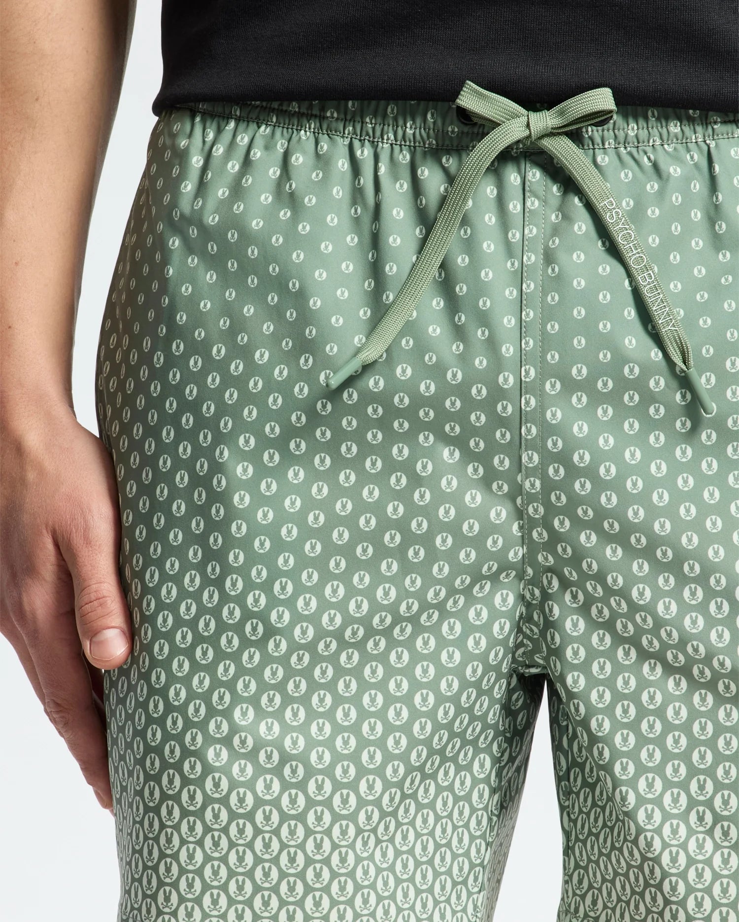 Someone is wearing the MENS BENDIGO PRINTED SWIM TRUNK by Psycho Bunny, which features a light green color with a small white pattern and an elasticized drawstring waist. The slightly glossy texture of the trunks complements their black top as their hand rests casually on the side.