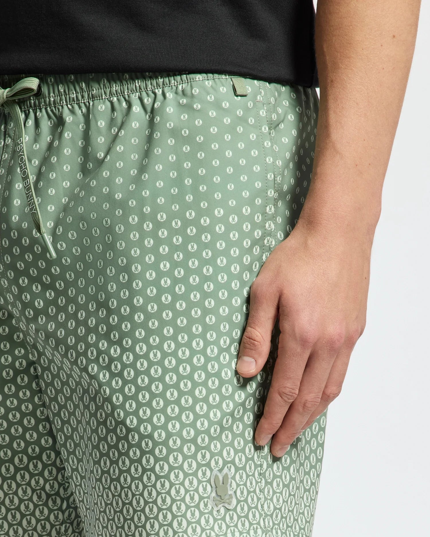 A person stands against a plain background wearing Psycho Bunny's MENS BENDIGO PRINTED SWIM TRUNK - B6W619F200, featuring a circular pattern and an elasticized waist with a black shirt. Part of their arm and hand is naturally visible by their side.