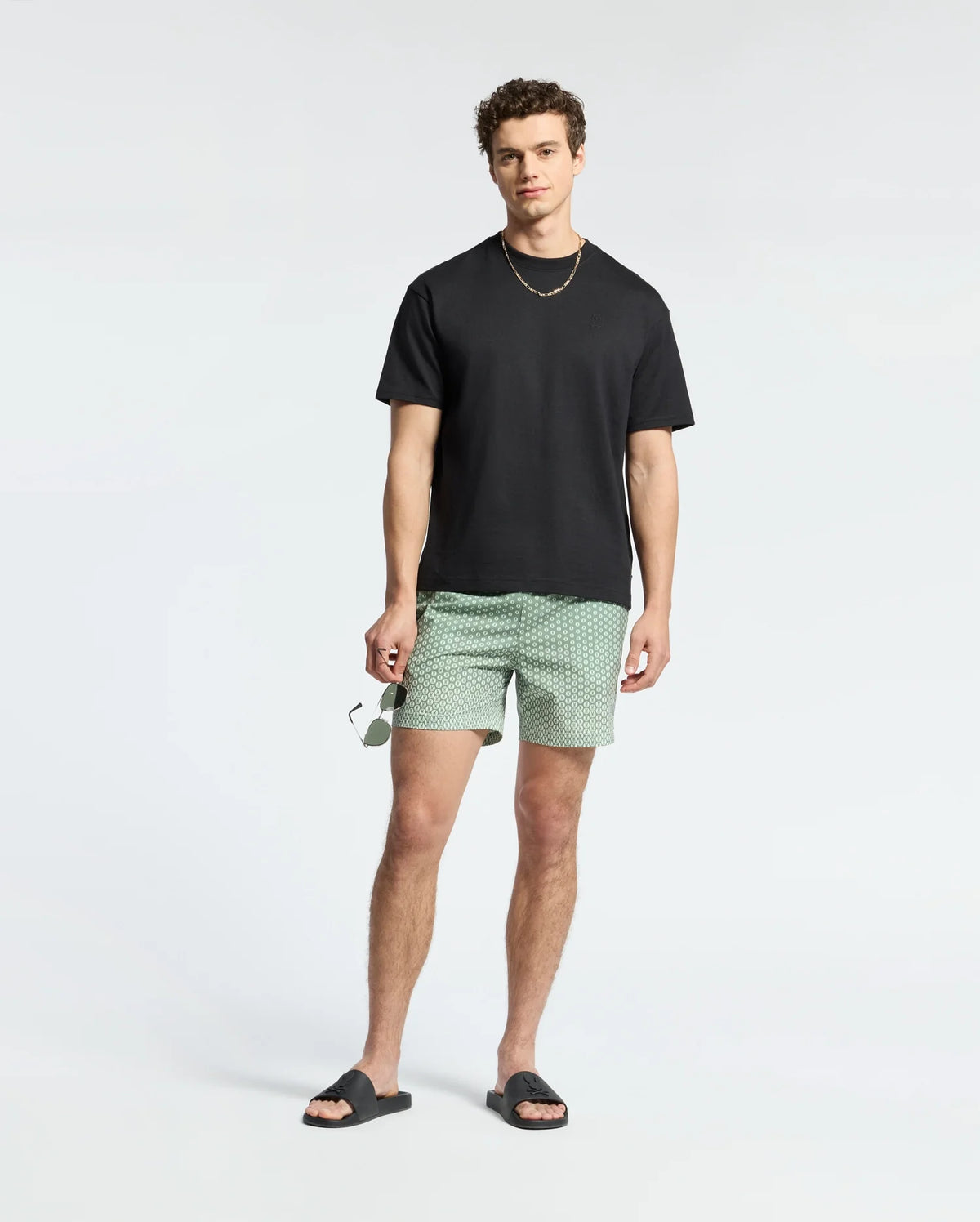 A man with curly hair stands against a plain background wearing a black T-shirt, Psycho Bunny's Bendigo printed swim trunks in green with an elasticized waist, black slide sandals, and a gold chain. He holds sunglasses in his right hand.