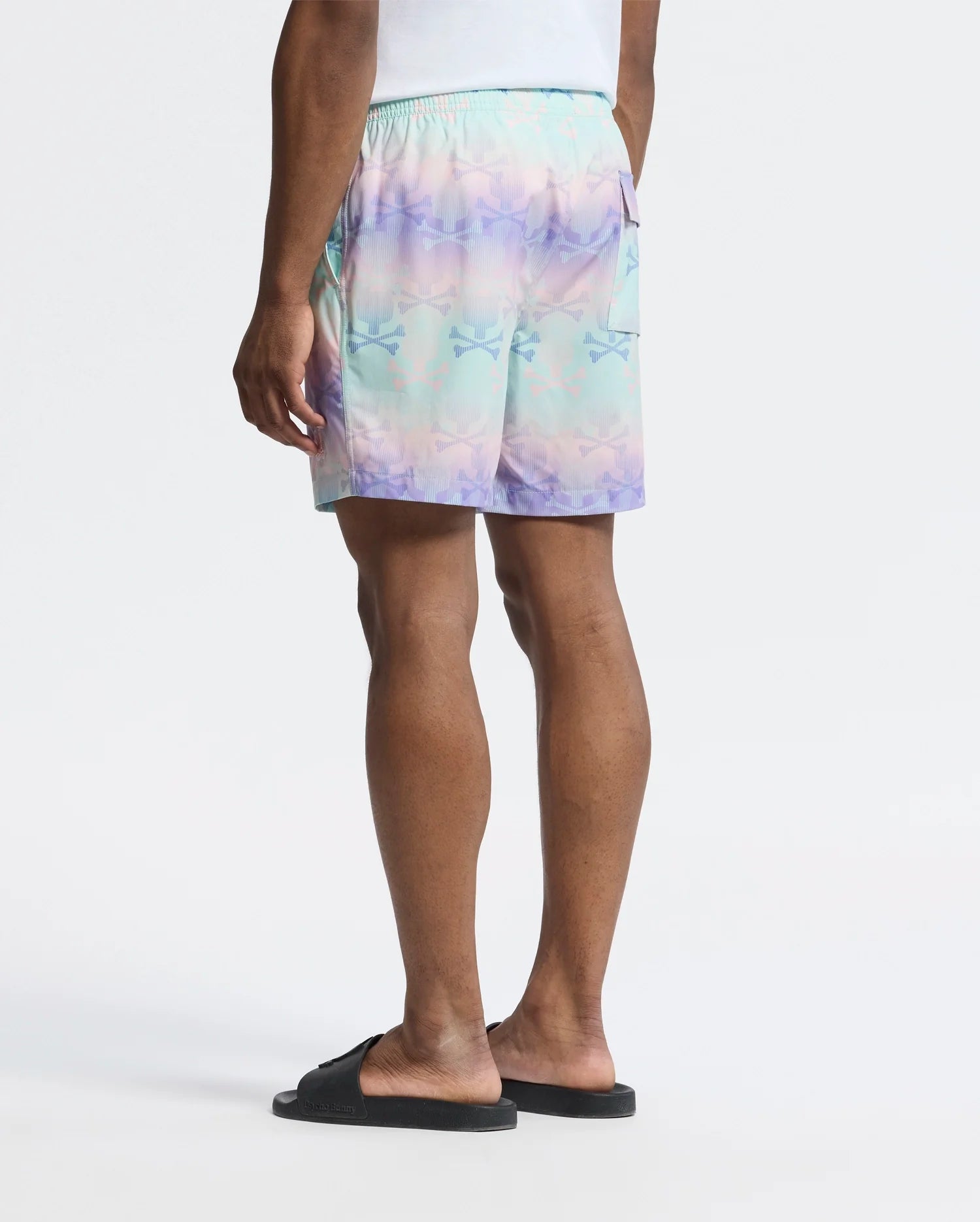Wearing Psycho Bunny's MENS FORSTER BUNNY PRINTED SWIM TRUNK, a person stands with back to the camera in pastel tie-dye shorts featuring a subtle Bunny print and black slides. The plain white backdrop emphasizes the relaxed, beach-bum vibe focused on their lower body.