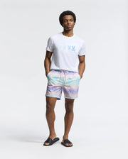A man exudes beach-bum comfort in a light T-shirt and Psycho Bunny pastel tie-dye MENS FORSTER BUNNY PRINTED SWIM TRUNK. With black slide sandals, he stands casually, hands in pockets, against a plain white background.