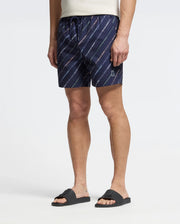 A person in a white t-shirt and Psycho Bunny's MENS RENMARK PB PRINTED SWIM TRUNK (B6W622F200) with diagonal stripes and logo print stands against a light background, paired with black sandals for a casual look.