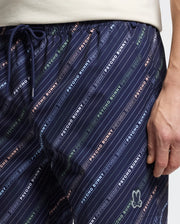 A close-up shows someone in the MENS RENMARK PB PRINTED SWIM TRUNK by Psycho Bunny. The dark blue trunks feature diagonal stripes, a colorful logo print, and a peace sign hand logo on the right leg, all set against a neutral backdrop.