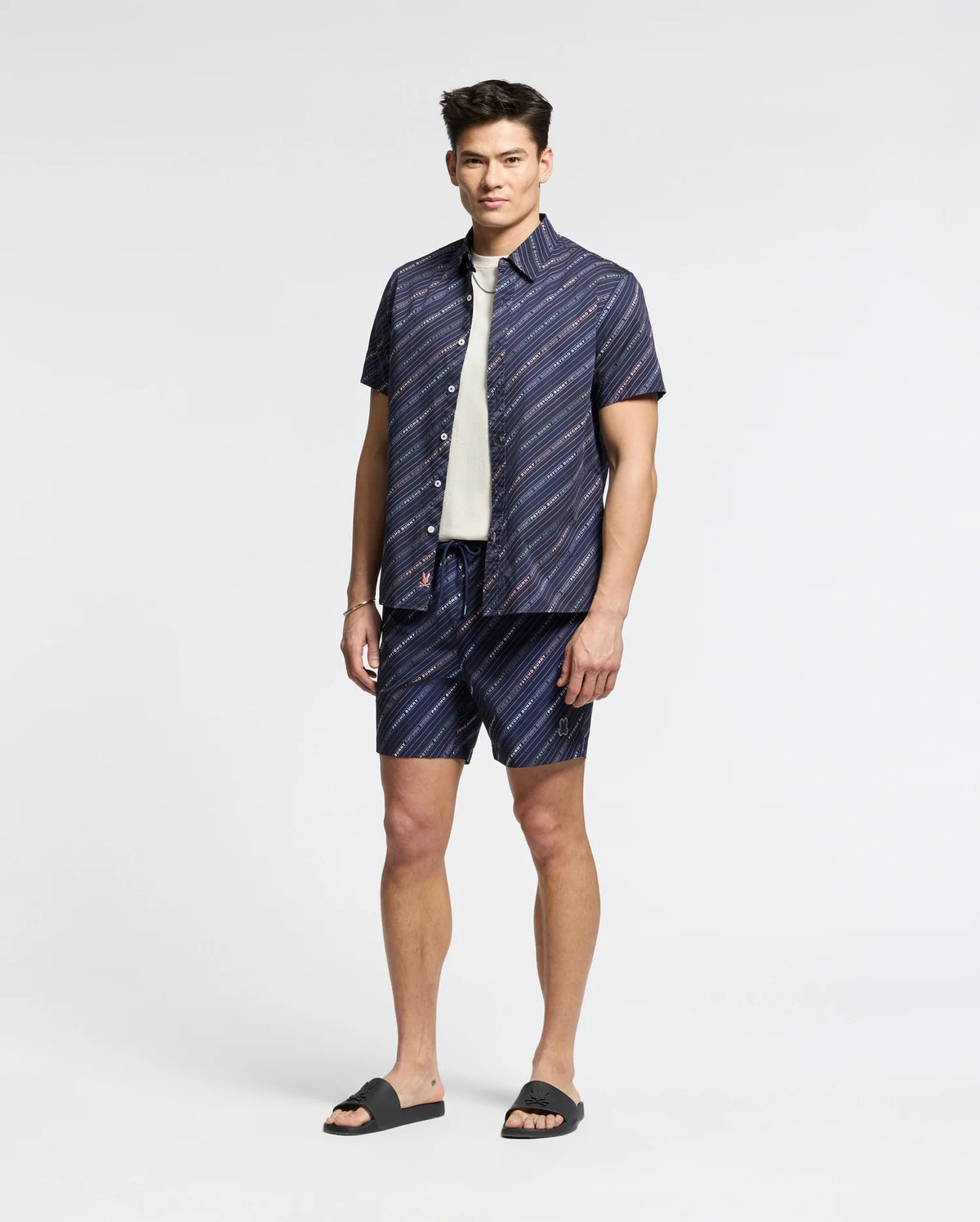 A man stands against a plain background in a relaxed pose, wearing Psycho Bunny’s dark blue and white striped MENS RENMARK PB PRINTED SWIM TRUNK (B6W622F200), paired with a white t-shirt and dark sandals, exuding style as if ready to toss essentials into his waterproof bag.