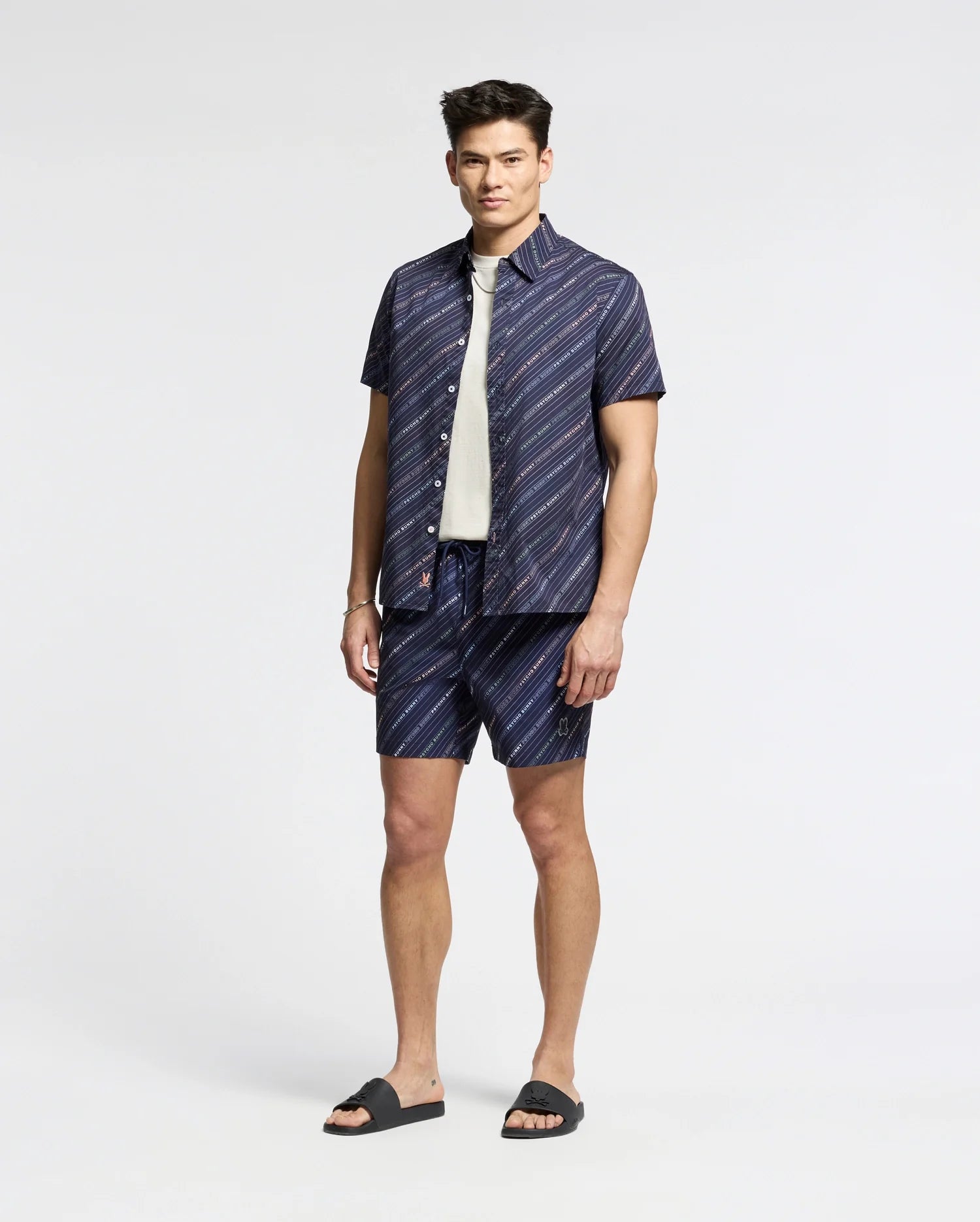 A man stands against a plain background in a relaxed pose, wearing Psycho Bunny’s dark blue and white striped MENS RENMARK PB PRINTED SWIM TRUNK (B6W622F200), paired with a white t-shirt and dark sandals, exuding style as if ready to toss essentials into his waterproof bag.