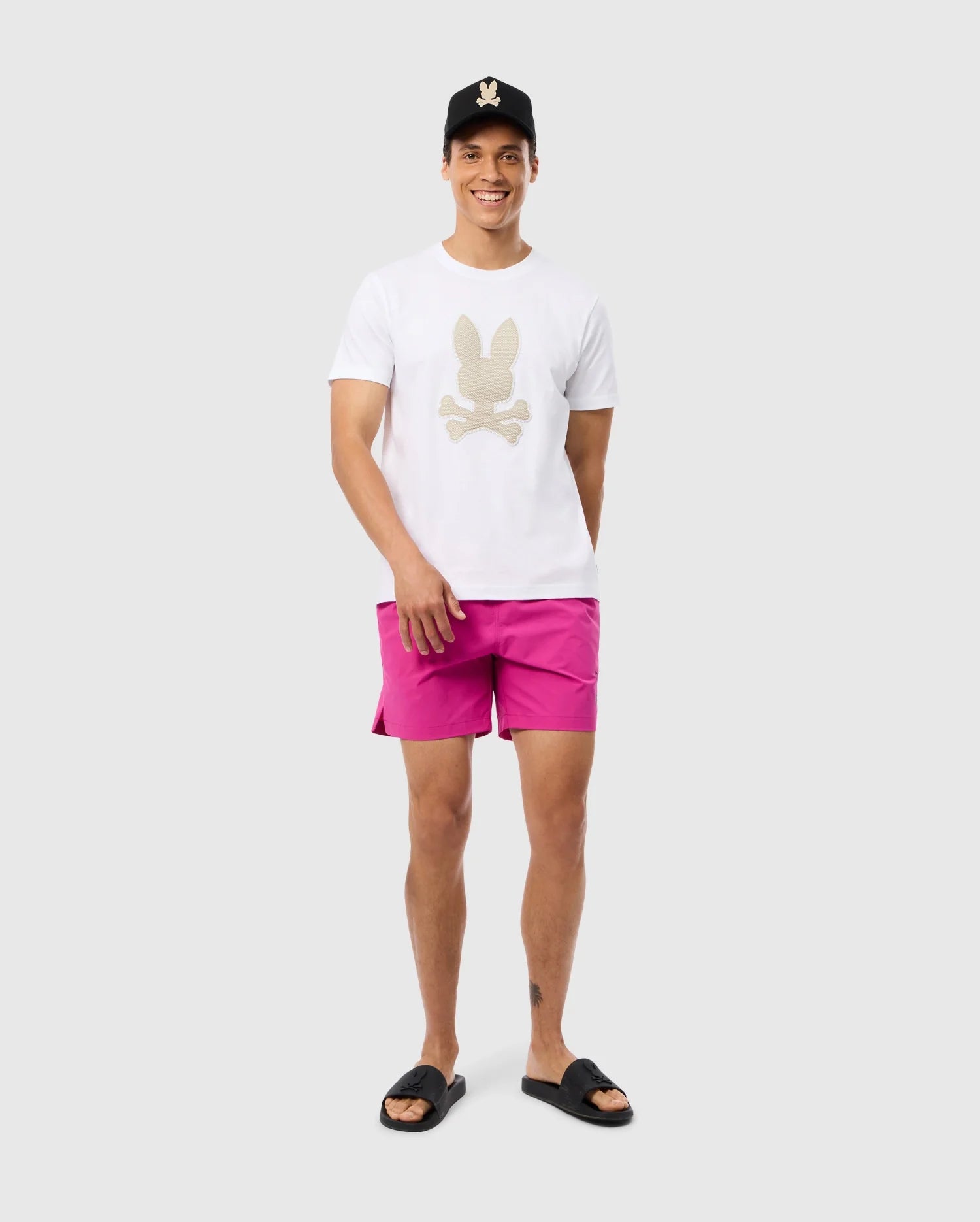 A person is standing and smiling while wearing a white t-shirt with a bunny pattern, the MENS PARKER HYDROCHROMIC SWIM TRUNK - B6W646C200 by Psycho Bunny, a black baseball cap, and black slide sandals. The background is plain and light-colored.