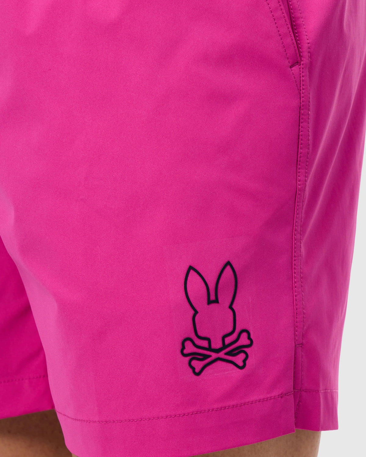 Close-up of a person wearing bright pink, quick-drying MENS PARKER HYDROCHROMIC SWIM TRUNK - B6W646C200 by Psycho Bunny with side pockets. The trunks feature a black outline design logo near the hem showing a rabbit's head above crossed bones, resembling a playful skull and crossbones motif that adds to the unique bunny pattern.
