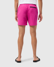 A person is standing with their back facing the camera, wearing bright pink Psycho Bunny MENS PARKER HYDROCHROMIC SWIM TRUNK - B6W646C200 with a small black pocket. Their white T-shirt and black flip-flops complete the look. The individual's arms are slightly bent, revealing tattoos against a plain white background.