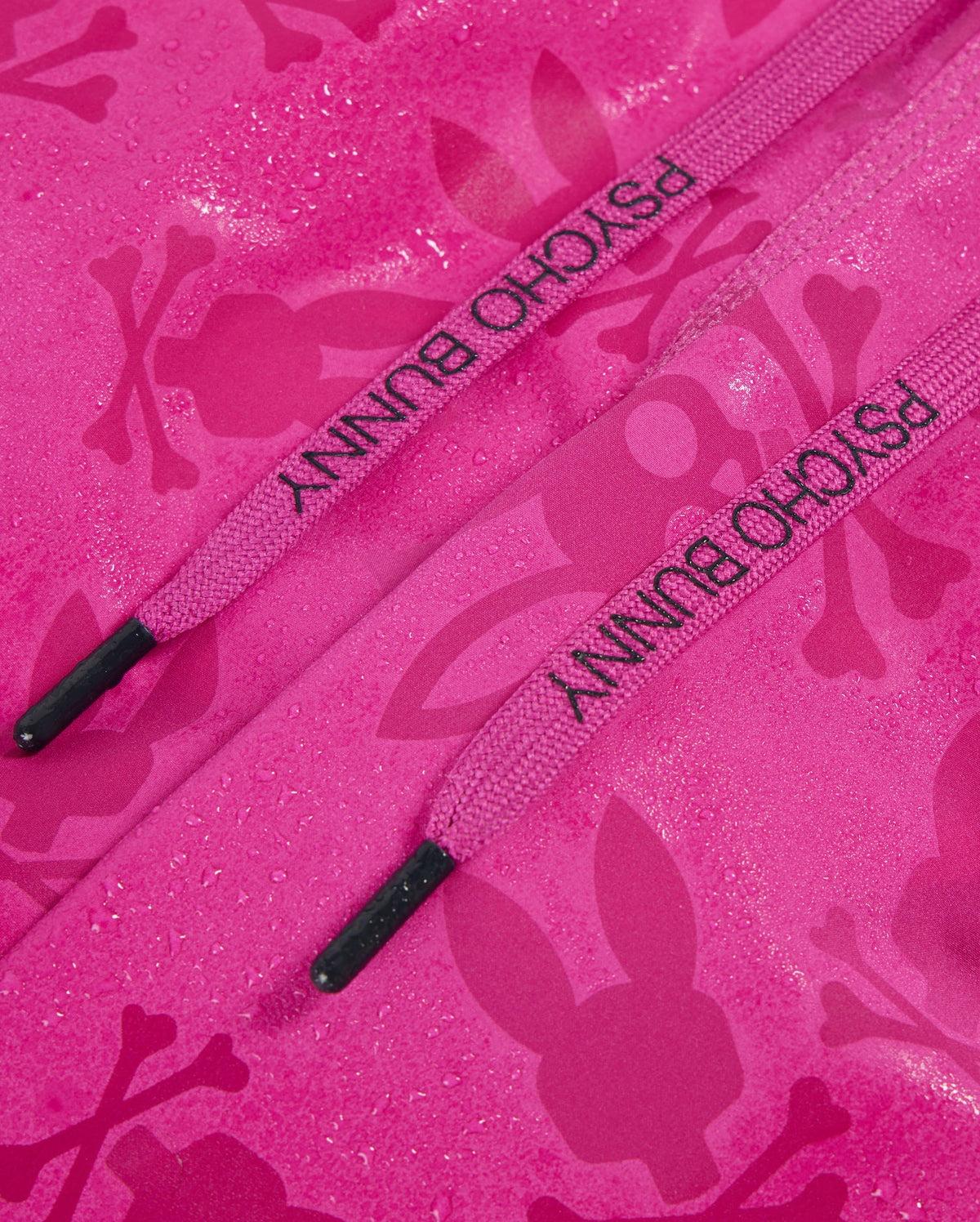 Close-up of a pink fabric with a bunny pattern and skull silhouettes. Two pink drawstrings with the text "PSYCHO BUNNY" rest against the quick-drying material, with droplets of water scattered on the surface, reminiscent of Psycho Bunny's MENS PARKER HYDROCHROMIC SWIM TRUNK - B6W646C200.