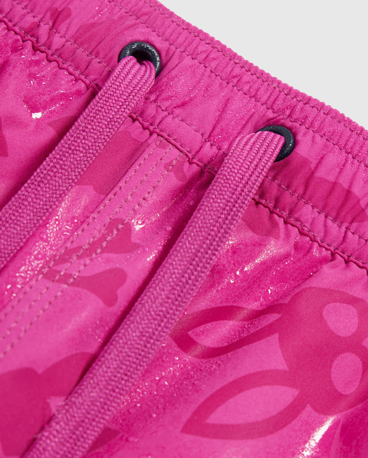 Close-up of bright pink Psycho Bunny MENS PARKER HYDROCHROMIC SWIM TRUNK - B6W646C200 featuring drawstrings and an elastic waistband. The fabric has a pattern with subtle skull and crossbones designs. The drawstrings are threaded through black metal eyelets, and the material appears to have a slight sheen.
