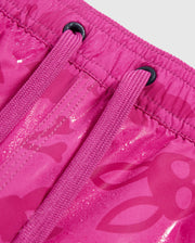 Close-up of bright pink Psycho Bunny MENS PARKER HYDROCHROMIC SWIM TRUNK - B6W646C200 featuring drawstrings and an elastic waistband. The fabric has a pattern with subtle skull and crossbones designs. The drawstrings are threaded through black metal eyelets, and the material appears to have a slight sheen.