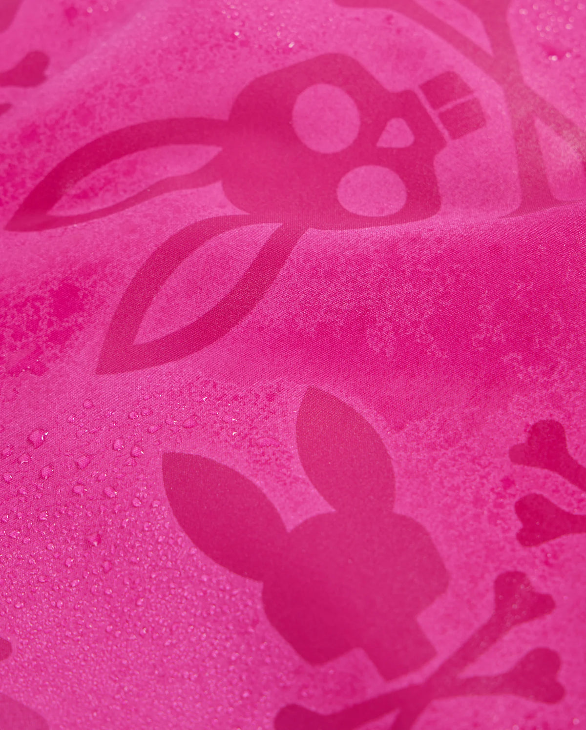 A close-up of quick-drying pink MENS PARKER HYDROCHROMIC SWIM TRUNK - B6W646C200 by Psycho Bunny, featuring a bunny pattern with skulls and crossbones. The swim trunk fabric appears to be slightly wet, with droplets of water visible on the surface. The design elements are in a darker shade of pink, creating a subtle contrast.
