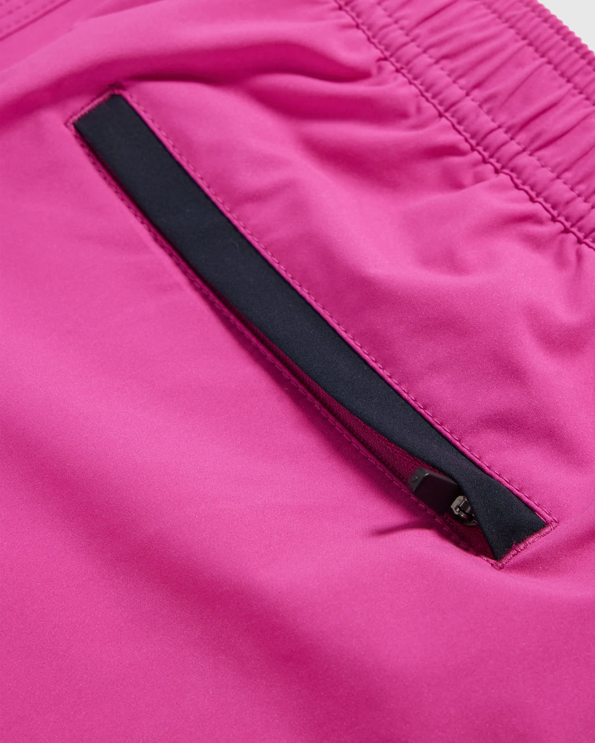 Close-up image of a bright pink fabric on quick-drying MENS PARKER HYDROCHROMIC SWIM TRUNK - B6W646C200 by Psycho Bunny, featuring a black zippered pocket. The pocket zipper is partially visible against the vibrant pink background. The fabric appears smooth and slightly shiny, suggesting a likely synthetic material.