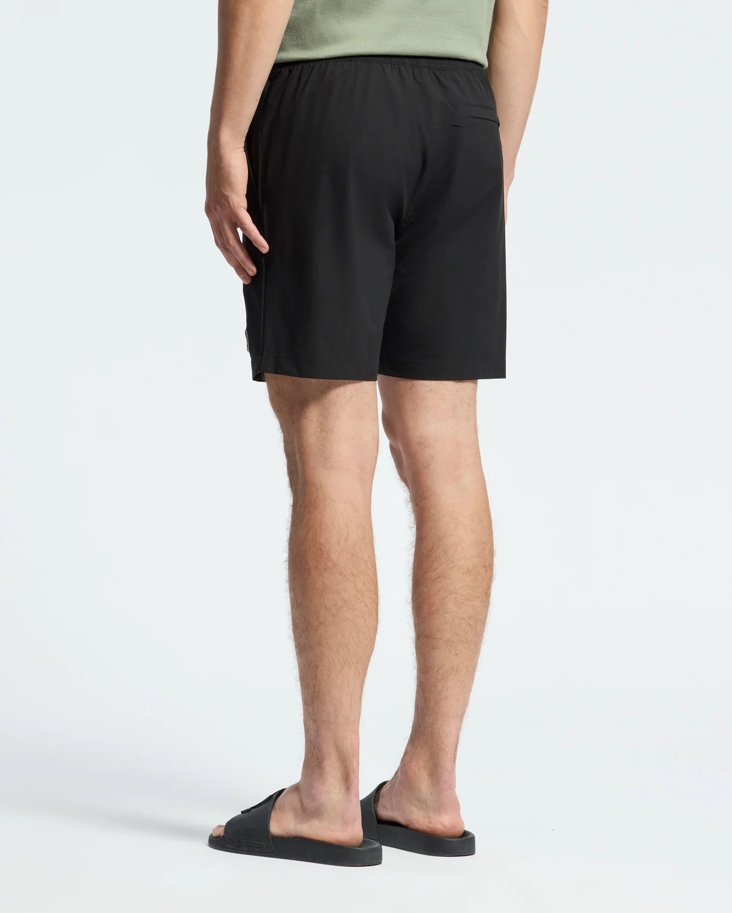 Rear view of a person in a light green T-shirt, Psycho Bunny MENS SAMOS 7 HYDROCHROMIC SWIM TRUNK, and black slip-on sandals, capturing the laid-back Sunsoaker style against a plain white background.
