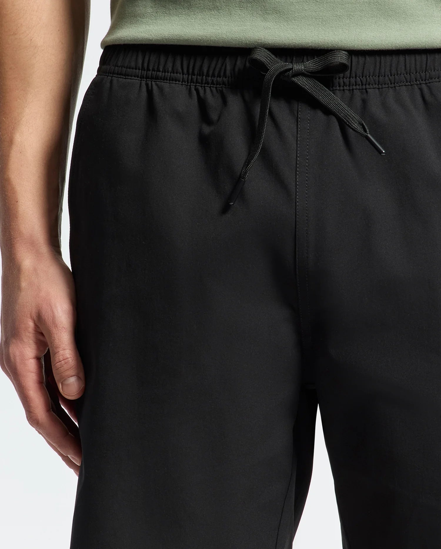 A person in black Psycho Bunny MENS SAMOS 7 HYDROCHROMIC SWIM TRUNK, featuring an elastic waistband and drawstring in a casual sunsoaker style. The top of a light green shirt is visible, and their slightly bent left arm rests alongside their body.