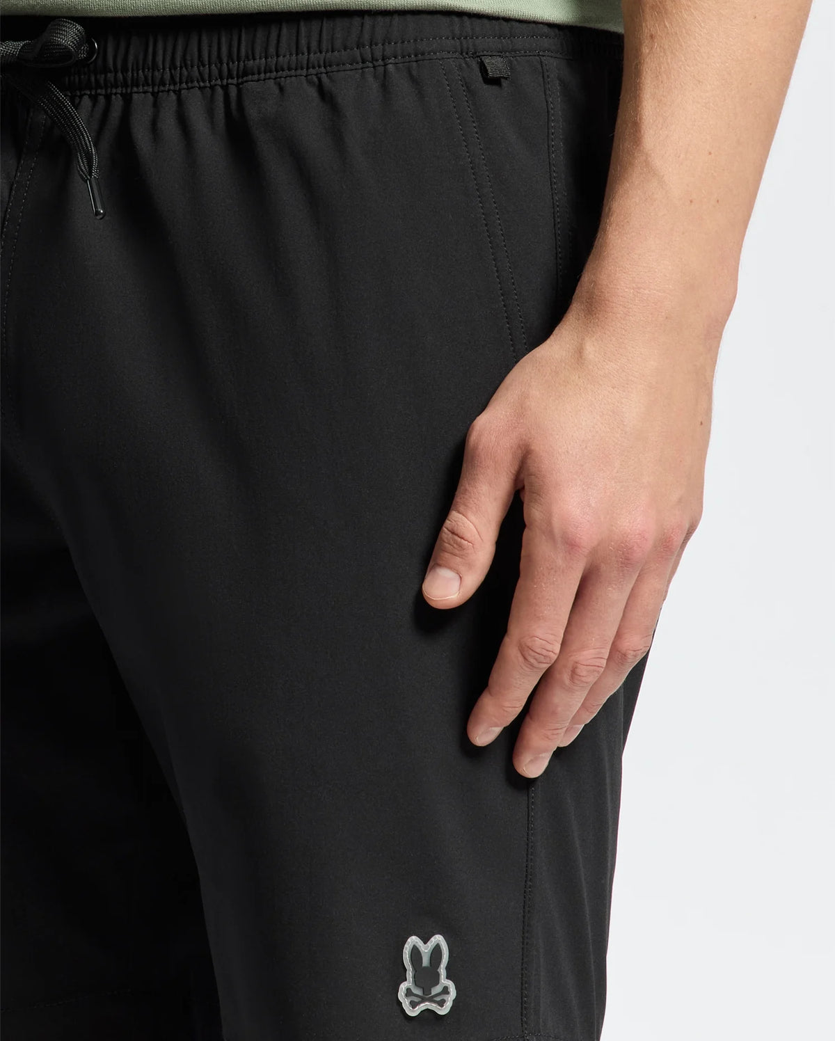 A person wearing Psycho Bunny's black MENS SAMOS 7 HYDROCHROMIC SWIM TRUNK - B6W650F200 with a small bunny logo on the left thigh stands casually, their right hand by their side. These sunsoaker style trunks feature a drawstring waist for an adjustable fit.