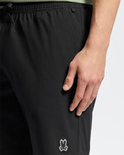 A person wearing Psycho Bunny's black MENS SAMOS 7 HYDROCHROMIC SWIM TRUNK - B6W650F200 with a small bunny logo on the left thigh stands casually, their right hand by their side. These sunsoaker style trunks feature a drawstring waist for an adjustable fit.