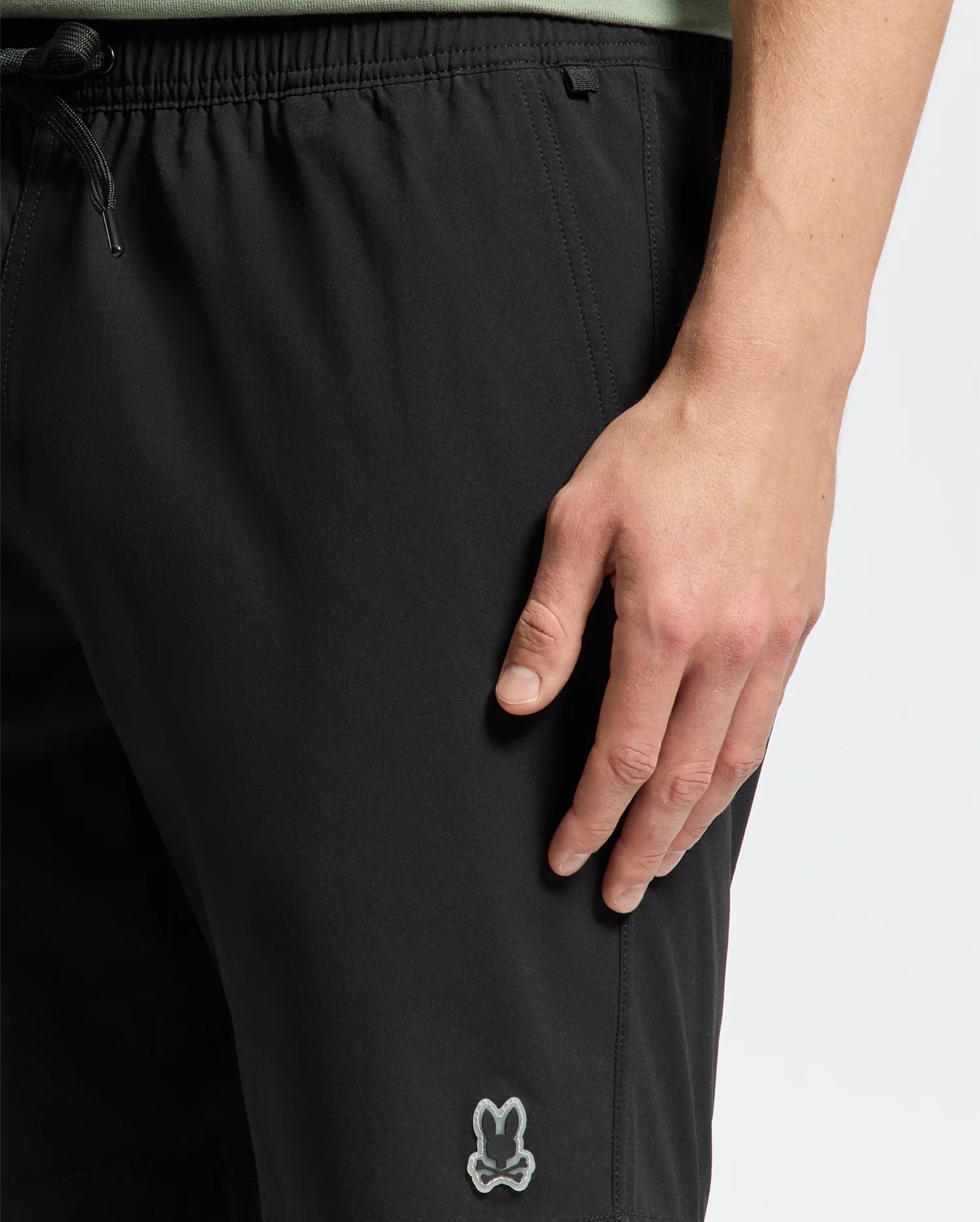 A person wearing Psycho Bunny's black MENS SAMOS 7 HYDROCHROMIC SWIM TRUNK - B6W650F200 with a small bunny logo on the left thigh stands casually, their right hand by their side. These sunsoaker style trunks feature a drawstring waist for an adjustable fit.