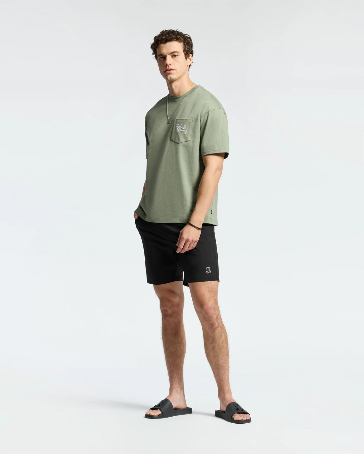 A man in a light green T-shirt with a pocket, wearing Psycho Bunny's MENS SAMOS 7 HYDROCHROMIC SWIM TRUNK and black sandals, stands against a plain background. Embracing the sunsoaker style, his hands rest in his pockets as he casually looks to the side.