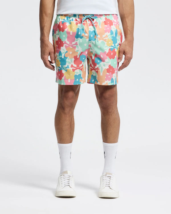 A man adorned in Psycho Bunny's Mens Lemmon All Over Print Swim Trunk (B6W658F200) is seen wearing colorful floral shorts in pink, yellow, blue, and peach hues. Paired with a white T-shirt, white socks, and sneakers, he stands against a plain backdrop.