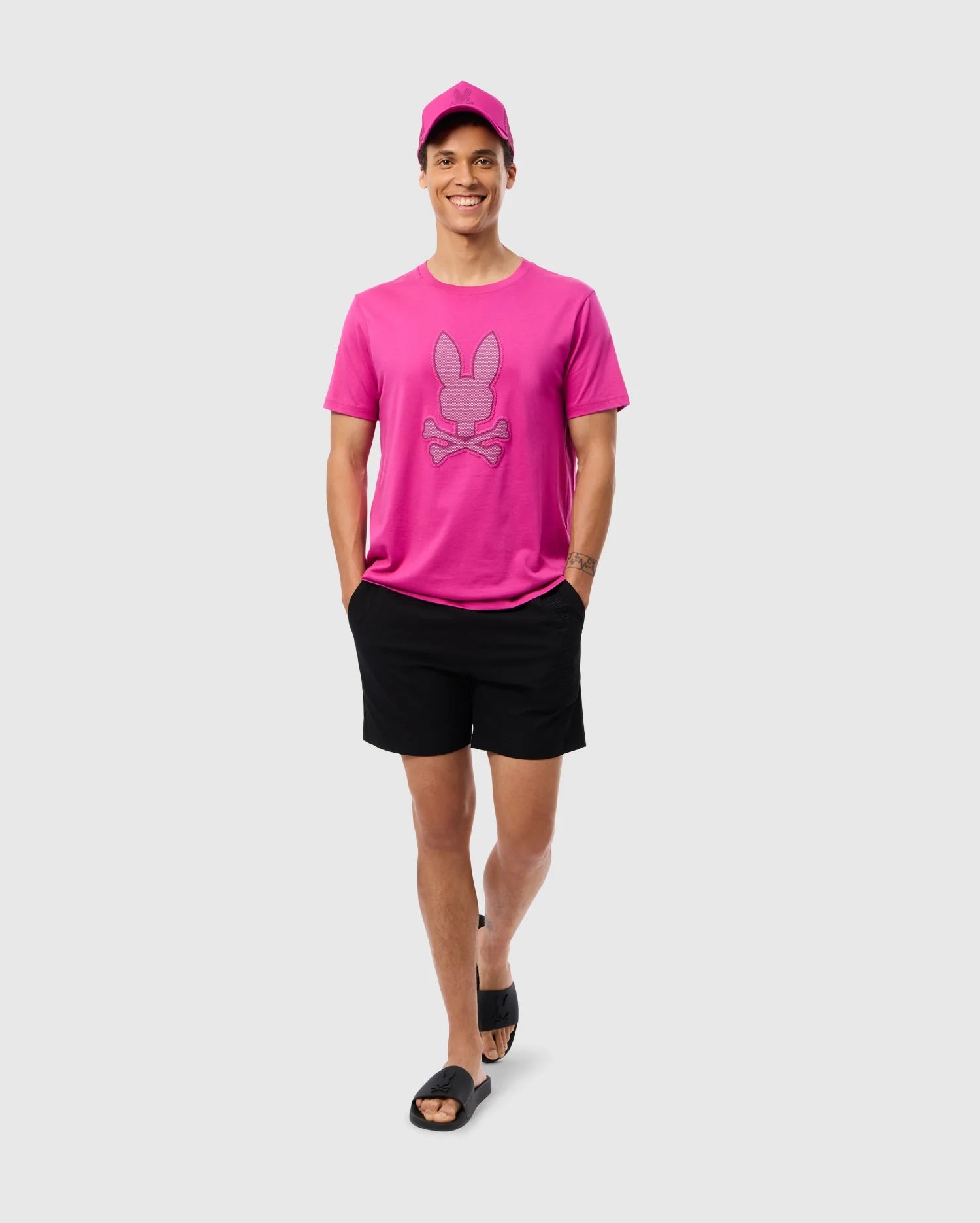 A smiling person is standing against a plain background, wearing a bright pink cap and matching pink t-shirt featuring a stylized bunny graphic. They are also wearing Psycho Bunny MENS MELVIN SWIM TRUNK - B6W665C200 and black slides, with hands in pockets, looking ready for summer fun.