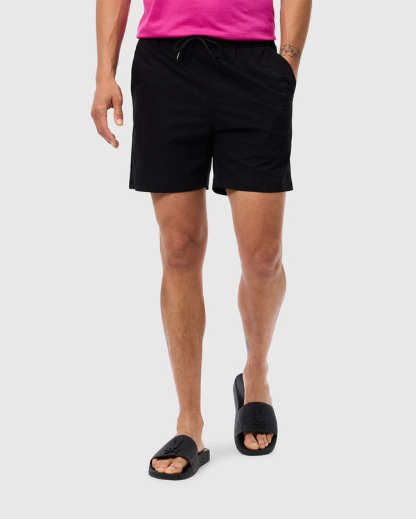 A person is wearing black Psycho Bunny MENS MELVIN SWIM TRUNK - B6W665C200 with an elastic waistband and a pink shirt that is partially visible. They are also sporting black slide sandals with a cross design on the straps. A waterproof bag hangs casually by their side against the plain gray background.