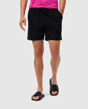 A person is wearing black Psycho Bunny MENS MELVIN SWIM TRUNK - B6W665C200 with an elastic waistband and a pink shirt that is partially visible. They are also sporting black slide sandals with a cross design on the straps. A waterproof bag hangs casually by their side against the plain gray background.