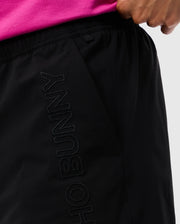 A person in a pink top places their hand in the pocket of black athletic swim trunks. The words "Psycho Bunny" are embroidered vertically in black thread along the side of these quick-dry shorts, identified as MENS MELVIN SWIM TRUNK - B6W665C200.