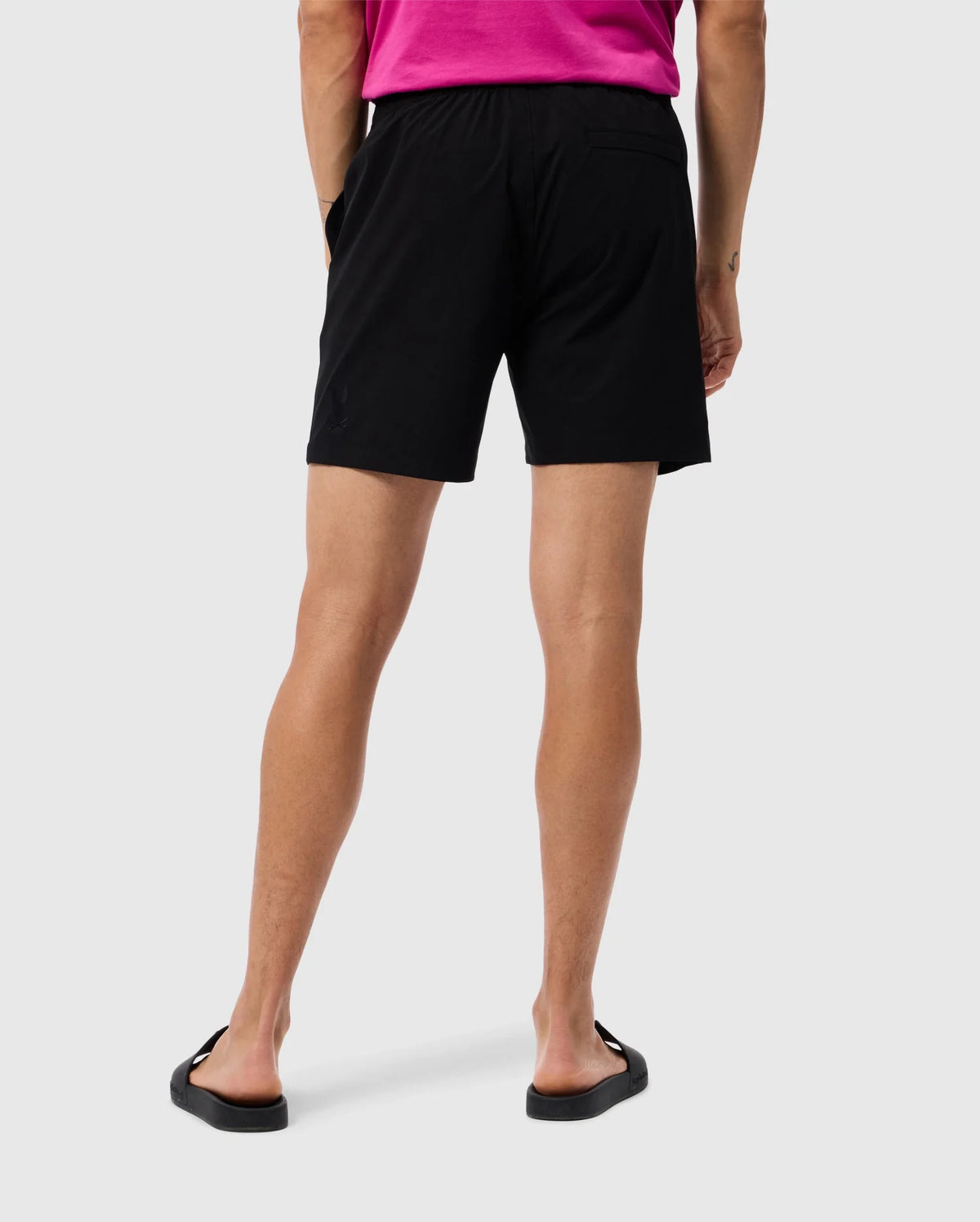 A person with light skin, wearing a bright pink shirt, Psycho Bunny MENS MELVIN SWIM TRUNK - B6W665C200, and black flip-flops, is standing with their back facing the camera. The background is plain white.